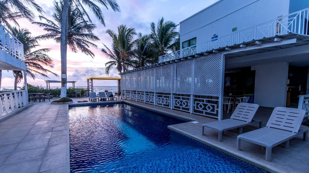 Área de piscina exterior en frente al mar con sillas reclinables, palmeras y cielo al atardecer
