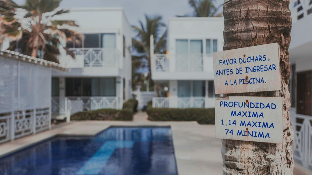 Piscina rodeada de edificios blancos, palmeras y carteles con normas de uso en español